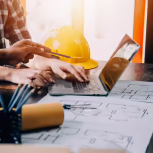 Two professionals reviewing construction blueprints and project plans on a laptop beside a yellow hard hat.